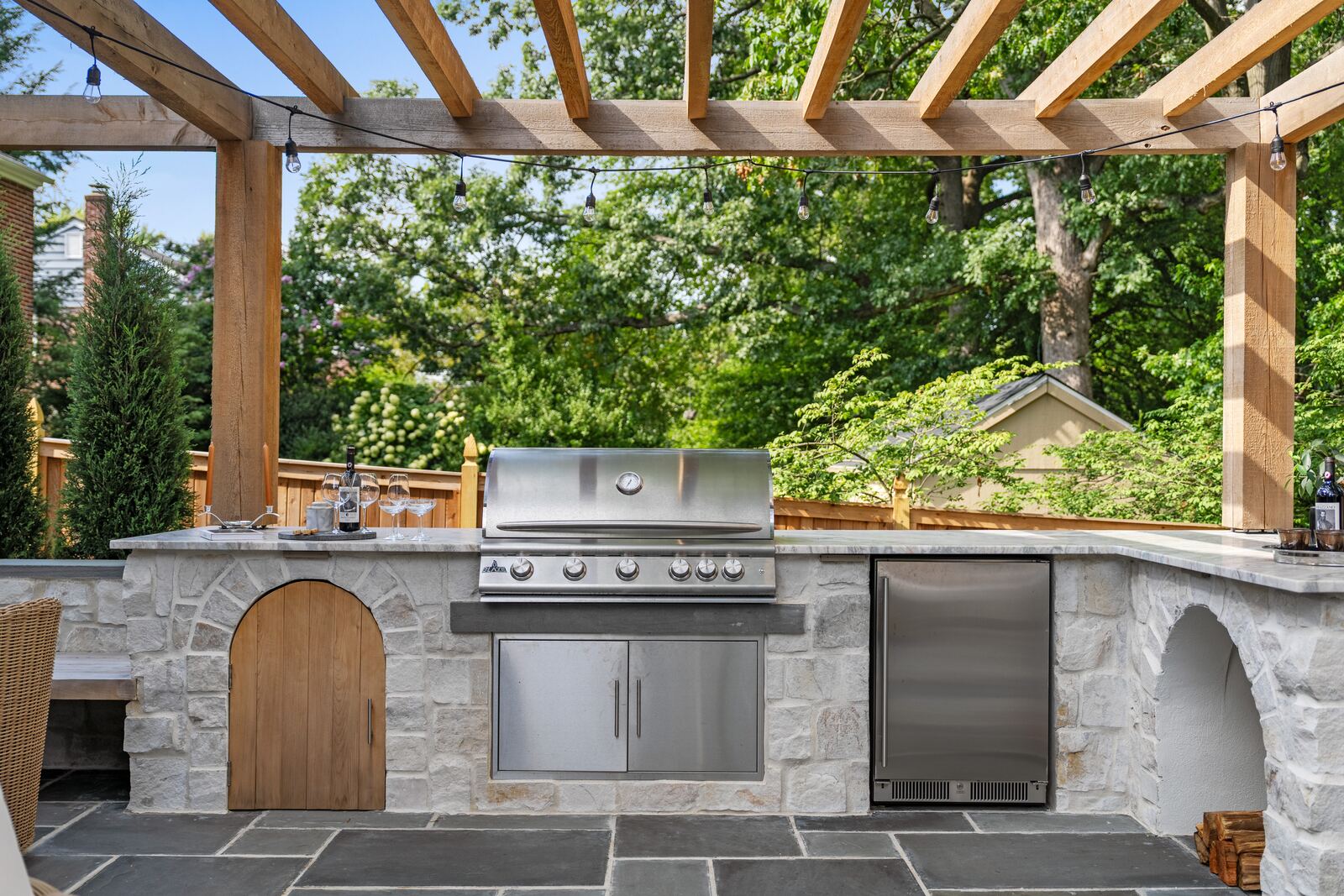 Stone outdoor kitchen with built-in grill and refrigerator under a cedar pergola