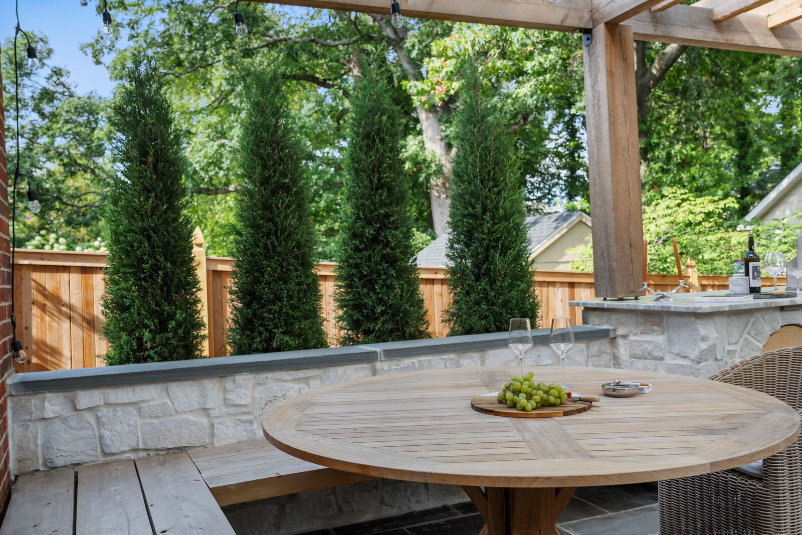 Stone seat wall around a teak dining table, backed by columnar trees and a cedar fence